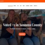 Homepage of K9 Activity Club & Lodge, featuring two smiling women sitting on a wooden bench with a Golden Doodle and a German Shepherd. The text overlay reads 'Voted #1 in Sonoma County' with a tagline 'The Premier Destination for Daycare, Training & Lodging.' The website's navigation bar includes links for services like daycare, training, and lodging, with a prominent 'Reservation' button. The background shows an outdoor setting with shaded seating and trees.