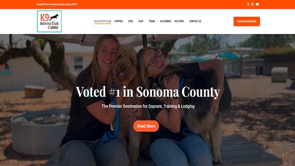 Homepage of K9 Activity Club & Lodge, featuring two smiling women sitting on a wooden bench with a Golden Doodle and a German Shepherd. The text overlay reads 'Voted #1 in Sonoma County' with a tagline 'The Premier Destination for Daycare, Training & Lodging.' The website's navigation bar includes links for services like daycare, training, and lodging, with a prominent 'Reservation' button. The background shows an outdoor setting with shaded seating and trees.