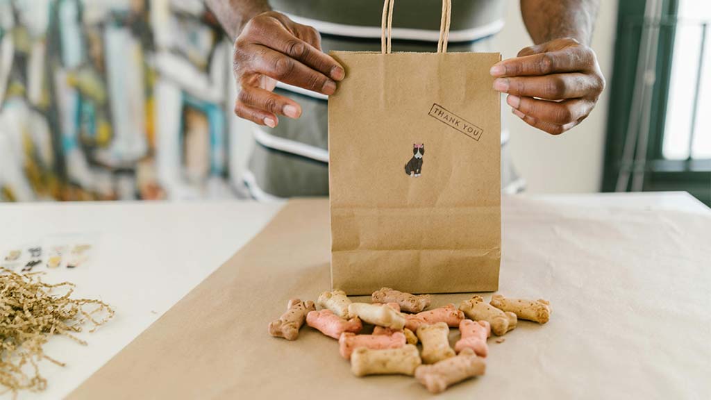 A man wearing an apron is packaging homemade dog treats into a brown paper bag labeled "Thank You," featuring a small dog sticker. The treats, shaped like bones, come in natural beige, pink, and light brown colors, suggesting organic ingredients. The scene is set on a craft-style workspace with a rustic aesthetic, enhancing the homemade appeal. This image represents Easy Organic Dog Treats Recipes, showcasing DIY, healthy pet snacks made with simple, wholesome ingredients.