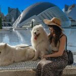 A woman wearing a straw hat, black tank top, and leopard-print skirt sits beside two fluffy white Samoyed dogs near a reflective water feature in a modern cityscape. The background showcases futuristic architecture, including curved glass structures and high-rise buildings under a clear blue sky. One dog is sitting upright, panting, while the other is lying down, looking relaxed. The scene captures a serene travel moment, highlighting the joy of exploring new places with pets. This image represents the experience of finding pet-friendly restaurants and activities while traveling, ensuring pets enjoy the journey as much as their owners.