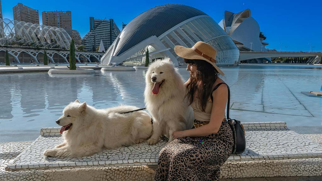 A woman wearing a straw hat, black tank top, and leopard-print skirt sits beside two fluffy white Samoyed dogs near a reflective water feature in a modern cityscape. The background showcases futuristic architecture, including curved glass structures and high-rise buildings under a clear blue sky. One dog is sitting upright, panting, while the other is lying down, looking relaxed. The scene captures a serene travel moment, highlighting the joy of exploring new places with pets. This image represents the experience of finding pet-friendly restaurants and activities while traveling, ensuring pets enjoy the journey as much as their owners.